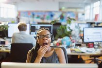 Mujer de negocios cansada con auriculares bostezando en la computadora en la oficina de planta abierta - foto de stock
