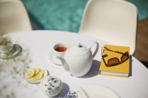 Serviço de chá e livro na mesa ensolarada à beira da piscina — Fotografia de Stock