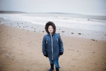 Retrato niño confiado en chaqueta de piel con capucha en la playa del océano invierno - foto de stock