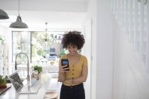 Sorridente giovane donna utilizzando smart phone in cucina — Foto stock