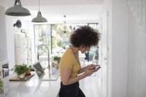 Giovane donna sms con smart phone in cucina — Foto stock