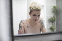 Portrait confident nude woman with tattooed shoulders at bathroom mirror — Stock Photo