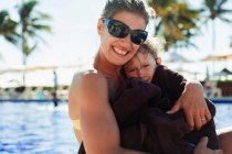 Ritratto madre tenuta figlio avvolto in asciugamano a bordo piscina soleggiata — Foto stock