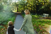 Feliz mãe e filho segurando as mãos e andando no parque de outono ensolarado — Fotografia de Stock