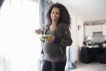 Retrato feliz joven embarazada comiendo ensalada en la ventana - foto de stock