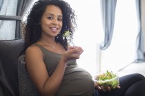 Retrato sonriente joven embarazada comiendo ensalada en el sofá - foto de stock