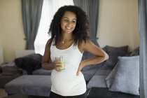 Glückliche junge schwangere Frau trinkt grünen Smoothie im Wohnzimmer — Stockfoto
