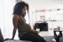 Mujer embarazada joven bebiendo batido verde en la sala de estar - foto de stock