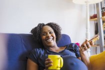 Risas de mujer embarazada con té y teléfono inteligente en el sofá - foto de stock