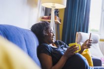 Mulher grávida feliz relaxando no sofá com chá e telefone inteligente — Fotografia de Stock