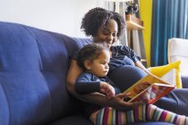Mãe grávida e filha lendo livro no sofá — Fotografia de Stock