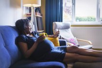 Щаслива вагітна жінка розслабляється на дивані з чаєм і смартфоном — стокове фото