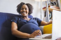 Feliz mujer embarazada que utiliza laptop en el sofá - foto de stock