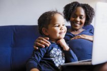Happy pregnant mother and preschool daughter using laptop on sofa — Stock Photo