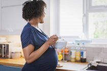 Femme enceinte mangeant et regardant la fenêtre de la cuisine — Photo de stock