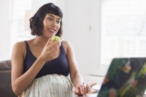 Счастливая беременная женщина ест тосты с авокадо за ноутбуком — стоковое фото