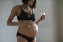 Беременная женщина в лифчике и трусиках наносит увлажняющий крем на живот — стоковое фото