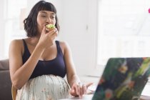 Mujer embarazada comiendo tostadas de aguacate en el portátil - foto de stock