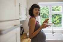 Portrait confident pregnant woman eating avocado toast in kitchen — Stock Photo