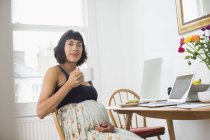 Portrait confident pregnant woman drinking tea at laptop — Stock Photo