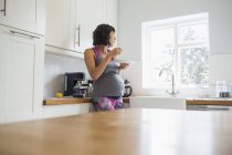 Задумчивая беременная женщина ест на кухне, глядя в окно — стоковое фото