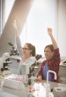 Eifrige Studentinnen heben im Labor-Klassenzimmer die Arme hinter Mikroskopen — Stockfoto