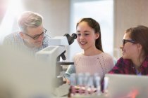 Male teacher and girl students conducting scientific experiment at microscope in laboratory classroom — Stock Photo
