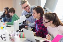 Улыбающийся учитель естественных наук помогает студенткам проводить научные эксперименты под микроскопом в лаборатории — стоковое фото