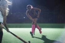 Determined young female field hockey player running with hockey stick, playing on field at night — Stock Photo