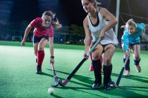 Young female field hockey players running to ball, playing field hockey on field at night — Stock Photo