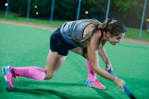 Determined young female field hockey player reaching with hockey stick, playing on field at night — Stock Photo