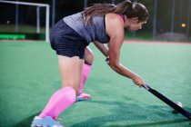 Determined young female field hockey player hitting the ball, playing on field at night — Stock Photo