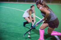 Determined young female field hockey players reaching for the ball with hockey sticks, playing on field — Stock Photo