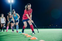 Determined young female field hockey player practicing sports drill on field at night — Stock Photo