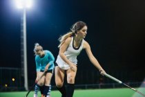 Focused young female field hockey player reaching with hockey stick, practicing on field at night — Stock Photo