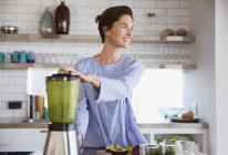 Smiling brunette woman making healthy green smoothie in blender in kitchen — Stock Photo