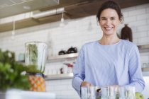Retrato sorrindo, mulher morena confiante preparando smoothie verde saudável no liquidificador na cozinha — Fotografia de Stock