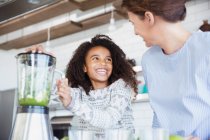 Энтузиастичная дочь помогает матери делать здоровый зеленый смузи в блендере на кухне — стоковое фото