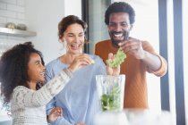 Familia multiétnica haciendo batido verde saludable en la licuadora en la cocina - foto de stock
