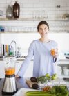 Портрет улыбается, уверенная брюнетка женщина пьет здоровый морковный сок в блендере на кухне — стоковое фото