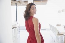 Porträt lächelnde, selbstbewusste brünette Frau im roten Kleid, die zu Hause über die Schulter schaut — Stockfoto