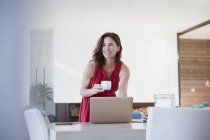Sorrindo morena mulher bebendo café, trabalhando no laptop na mesa da sala de jantar — Fotografia de Stock