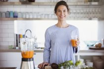 Retrato confiado, mujer sonriente beber jugo de zanahoria saludable en la cocina - foto de stock