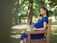 Беременная женщина разговаривает по мобильному телефону на скамейке в парке — стоковое фото