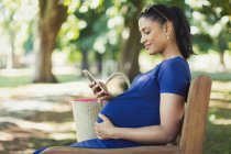 Pregnant woman texting with cell phone on park bench — Stock Photo