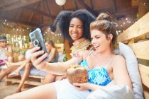 Young women friends with camera phone taking selfie drinking coconut cocktail at summer poolside — Stock Photo