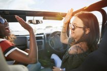 Jóvenes amigas chocando los cinco años en un jeep soleado, disfrutando de un viaje por carretera - foto de stock