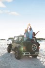 Exuberant young friends cheering with arms raised in jeep on beach, enjoying road trip — Stock Photo