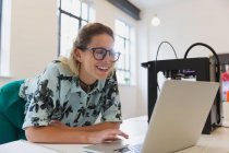 Усміхнена жінка-дизайнер, що працює в ноутбуці поруч з 3D принтером в офісі — стокове фото