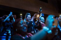 Sorrindo, público entusiasmado usando telefones de câmera no auditório escuro — Fotografia de Stock